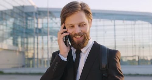 Hombre con barba que habla por teléfono cerca de la gran construcción de vidrio de la ciudad moderna