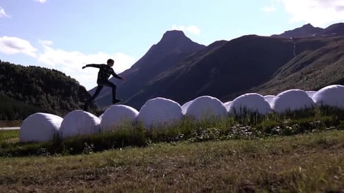 Slow motion shot of man jumping over bales of hay on a beautiful day with mountain background.