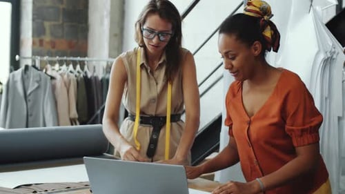 Two Multiethnic Seamstresses Chatting and Using Laptop in Sewing Studio