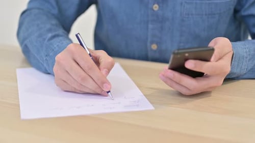 Nahaufnahme eines Mannes, der mit dem Smartphone auf Papier schreibt