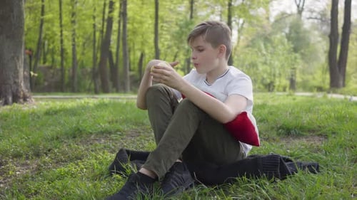 Young Boy Sit with Pillow in the Green Park and Eating an Apple. Outdoor Recreation