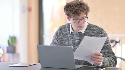 Young Adult Reviews Documents on Laptop in Office