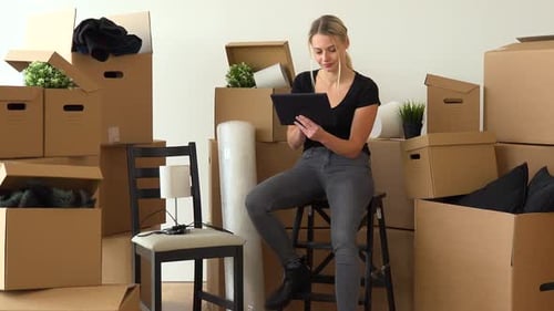 A Happy Moving Woman Sits on a Chair in an Empty Apartment and Works on a Tablet with a Smile