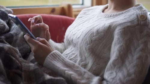 Nahaufnahme von Frauenhänden mit Smartphone zu Hause auf einem bequemen Stuhl in der Nähe des Fensters