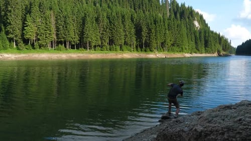 Young man throws the stone in the water of a mountain lake in the forest.