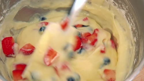 A Woman Stirs Yellow Cream with Fruits in a Steel Bowl with a Spatula - Closeup From Above