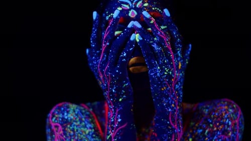 Woman's Face Painted with Fluorescent Paints Slowly Lowers Hands From Face Down Dark Background