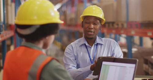 African american male and female workers wearing helmet and using laptop in warehouse