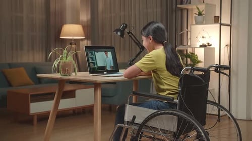 Asian Kid Girl Sitting In A Wheelchair While Learning Online With Teacher On Laptop Computer At Home