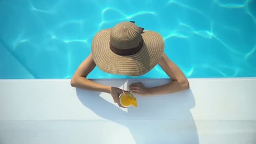 Young Woman in Elegant Hat Swimming in Pool and Enjoying Delicious Cocktail
