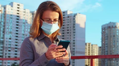 Portrait of a young woman in a protective mask using a mobile phone on the street.
