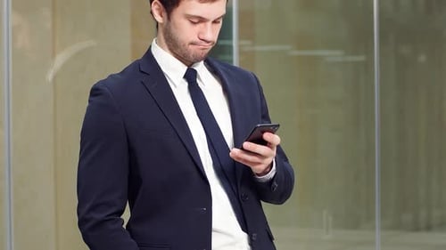 Portrait of Young Successful Businessman Using Smartphone and Looking at Camera in Modern Office