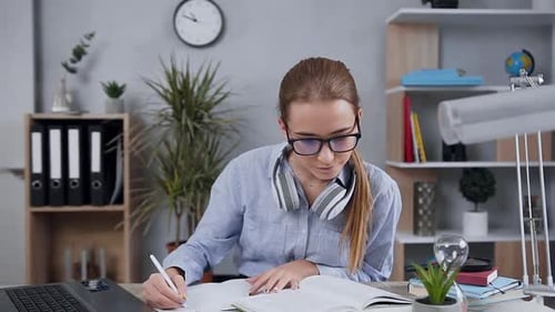 Blondes attraktives Mädchen mit Brille sitzt am Tisch und schreibt Essay mit Buch
