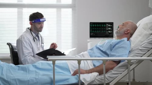 Young Doctor in Protective Mask Screen Talking to Senior Man Resting in Hospital Bed