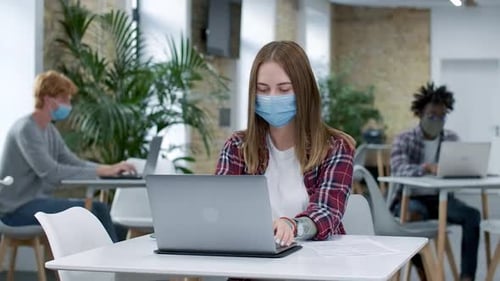 Young Focused Caucasian Woman in Covid-19 Face Mask Messaging Online in Office with Multiethnic