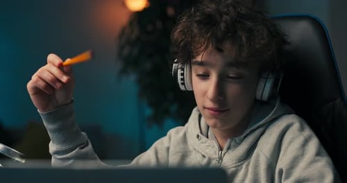 A Boy with Curly Hair Sits in a Room at a Desk with a Laptop on Wireless Headphones Taking Notes