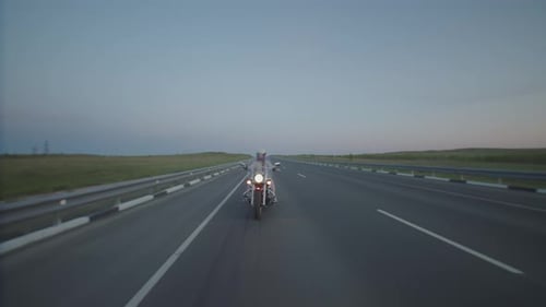 Woman rides forward on motorcycle on highway through countryside in evening, front view.