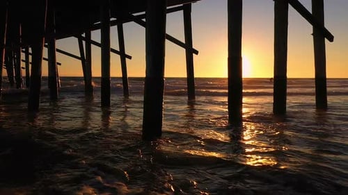 Panning view of sunset under pier as waves roll into the beach
