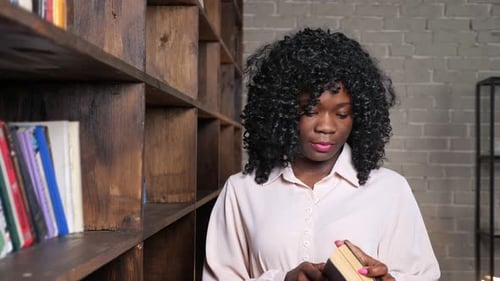Cheerful AfricanAmerican Woman Takes Book From Wooden Shelf