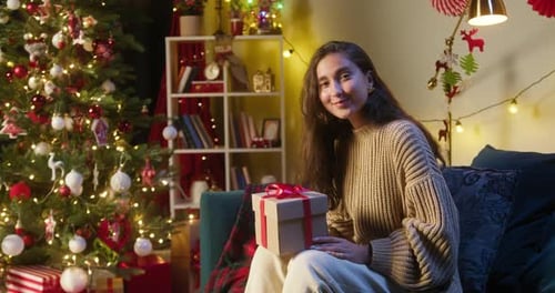 Woman Smiles with Christmas Gift by Decorated Tree