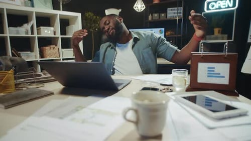 Portrait of Relaxed African American Guy Dancing Near Laptop at Workplace.