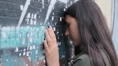 Sad Young Brunette Girl Stands with Her Hand Down and Leaning Her Head on the Wall with Graffiti