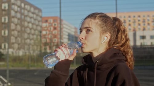 Young Woman Sits on Bench in Apartment Complex in Morning After Jogging and Drinks Water
