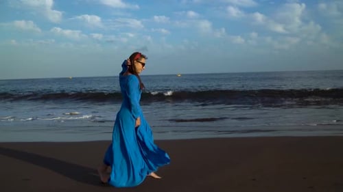 Frau in einem blauen Kleid läuft an einem schwarzen Vulkanstrand entlang