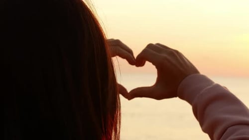Una mujer hermosa hace un corazón con sus manos al amanecer. La niña se regocija al amanecer