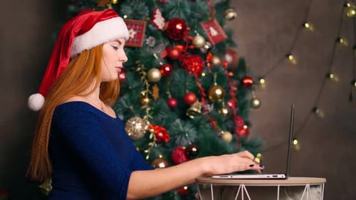 Frau in Weihnachtsmütze arbeitet Laptop und Typen auf Tastatur Hintergrund Weihnachtsbaum.