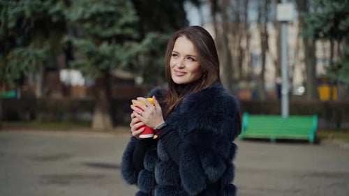 Positive young woman in the park. Attractive girl in stylish fur coat holds plastic cup of coffee an