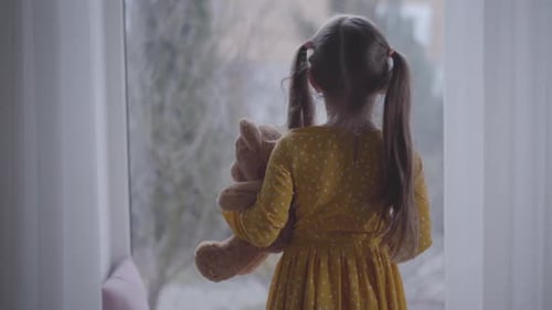Back View of Little Caucasian Girl Hugging Teddy Bear and Looking Out the Window. Brunette Kid