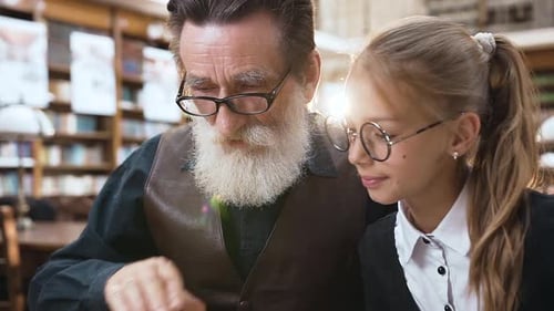 Old Grandpa Reading Interesting Book with His Smiling Teen Granddaughter in the Library