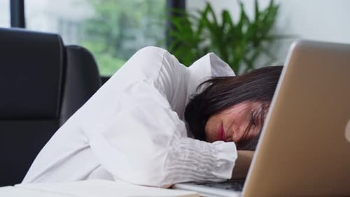 Asian young worker woman sleep on computer desk, feeling unwell and sick while working in office.