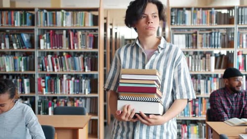 Serious Teenage Boy Walking in Library with Bunch of Books Looking Around