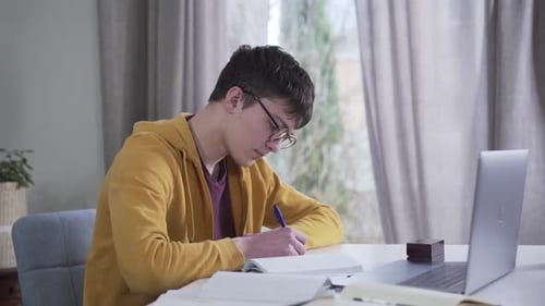Focused Caucasian Teenager in Eyeglasses Doing Homework Indoors. Smart Nale College Student Reading