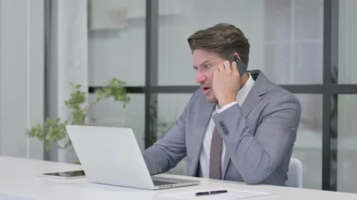 Angry Middle Aged Man Talking on Phone while using Laptop in Office
