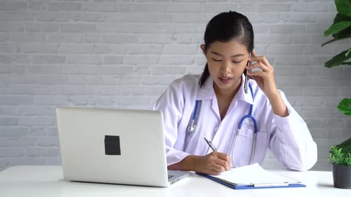 Concentrated Asian Female Doctor Talking on Phone While Using a Laptop at Doctor Office