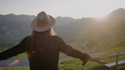 Rückansicht einer Hipster-Frau mit Hut im Licht einer Sonne, die ihre Hände gegen den Berg hebt