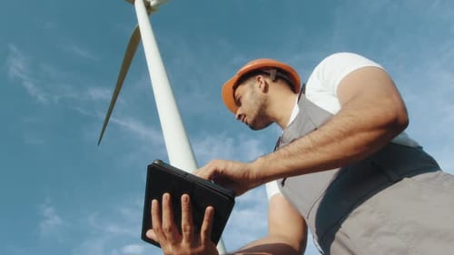 Indischer Ingenieur in Helm und grauem Overall steht zwischen Windmühlenfarm mit