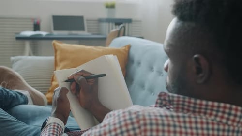 Man Lying on Sofa Writing in Notebook