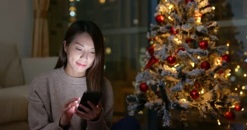 Woman Looks at Phone Next to Christmas Tree