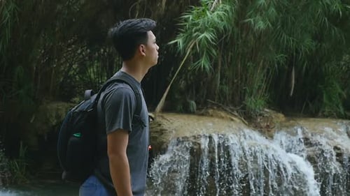 Young Man with Backpack Looks at Waterfall