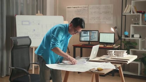 Asian Man Engineer With A Laptop Looking At Blueprint While Working At The Office