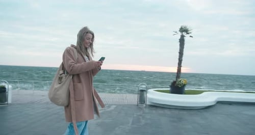 Young Woman Strolls Along Embankment and Chats Using Phone