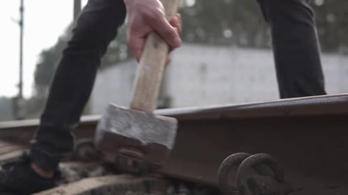 A railway man in a helmet repairs a railway, hammering the spire with a sledgehammer.