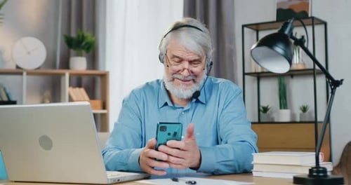 Älter Mann benutzt Handy, während er mit Laptop am Schreibtisch sitzt