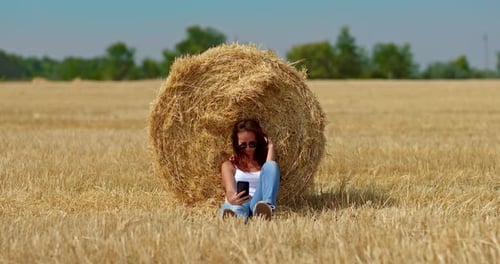 Young Woman Sits By a Large Golden Haystack in a Wheat Field and Takes a Selfie on Her Mobile Phone