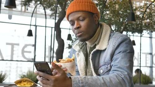 Young Adult Eating Burger and Using Smartphone
