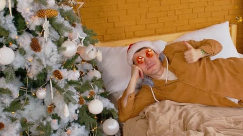 Jeune homme en bonnet de Père Noël et lunettes à la mode couché sur le lit près de l'arbre de Noël